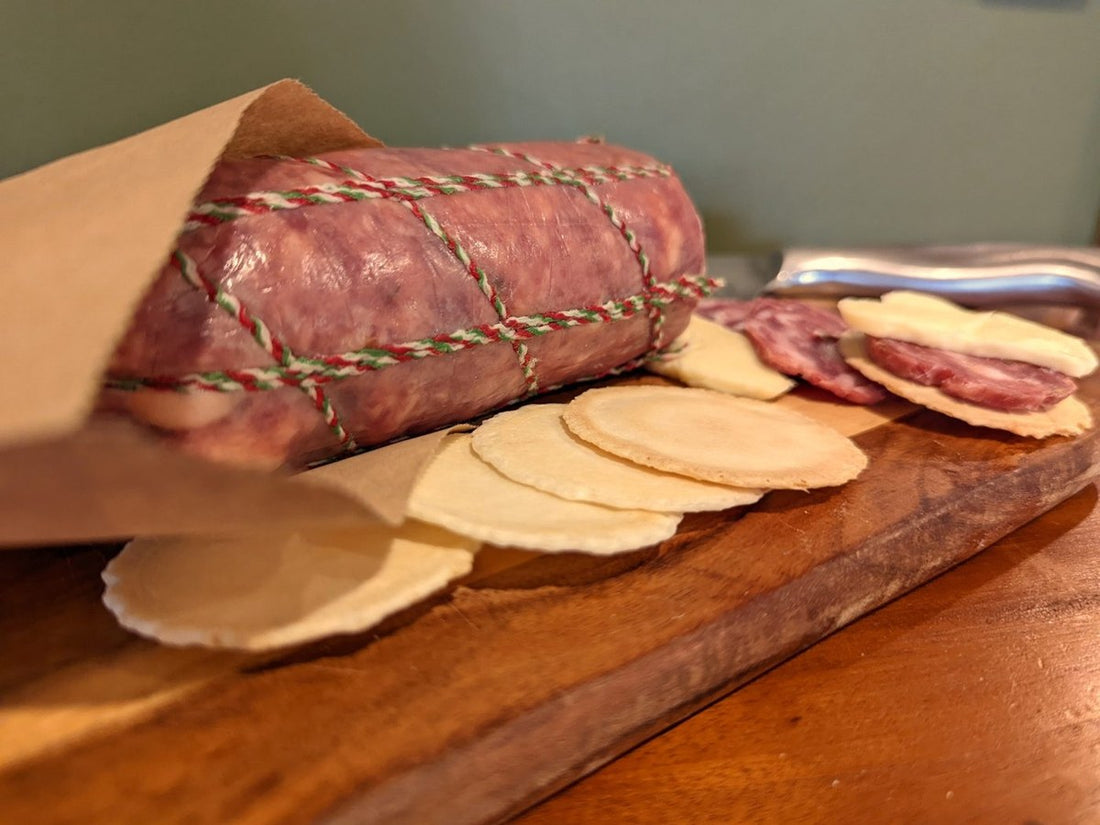 Whole salami wrapped in brown paper on a timber board with sliced salami and crackers