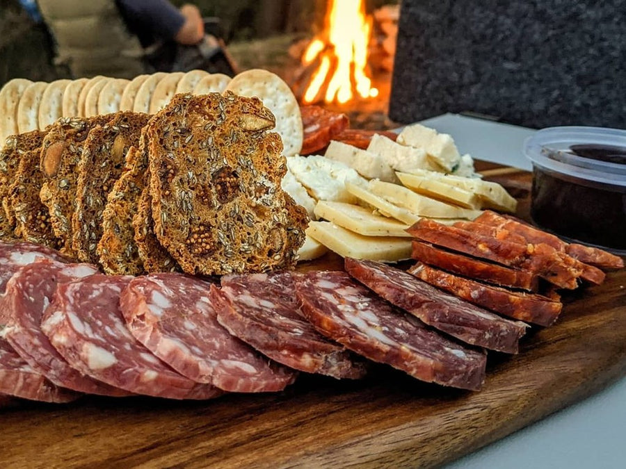 Sliced salami, cheese and crackers on a timber board beside a campfire