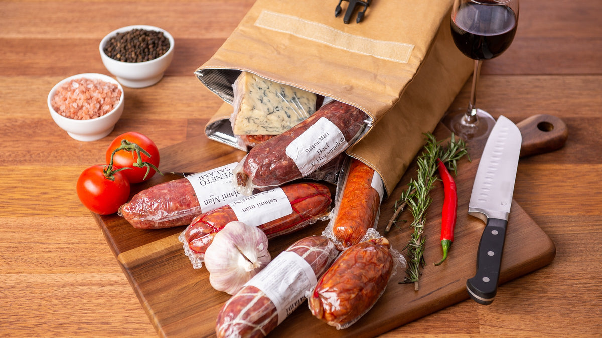 Vacuum-sealed Salami Man salami spilling from a bag onto a timber board with tomatoes, garlic, chilli and rosemary