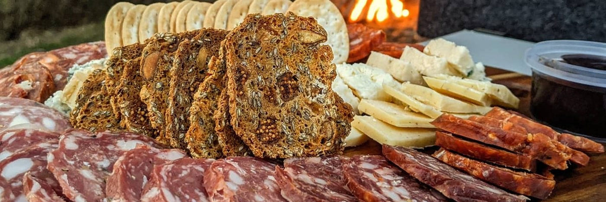Sliced salami, cheese and seeded crackers on a timber board by a campfire