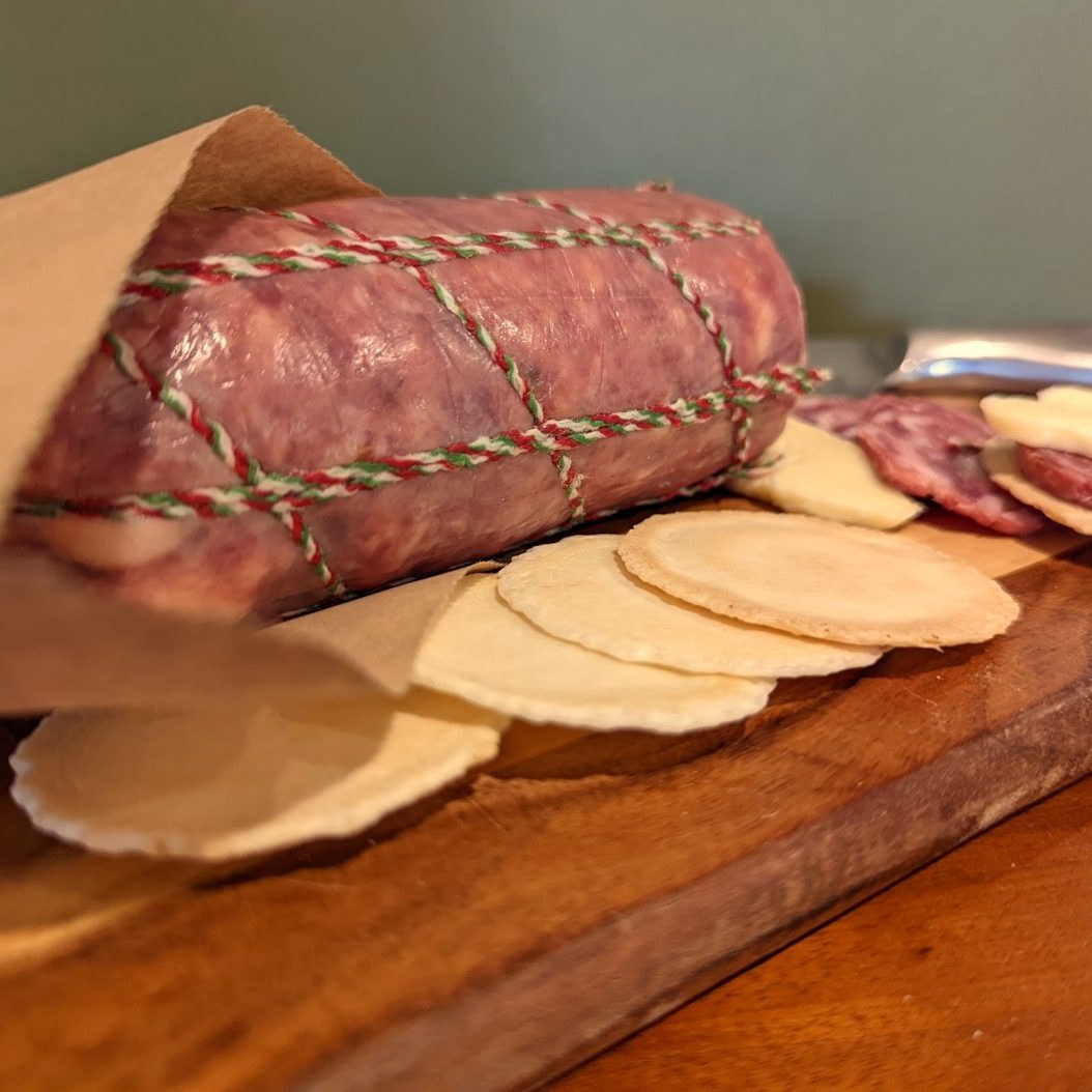 Whole salami wrapped in brown paper on a timber board with crackers and sliced salami