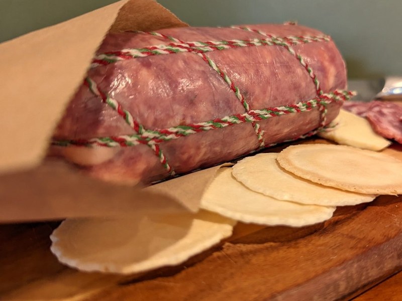 Whole salami tied with twine, wrapped in a brown paper bag on a timber board